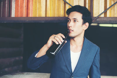 Young man drinking beer from bottle while standing against patterned metal