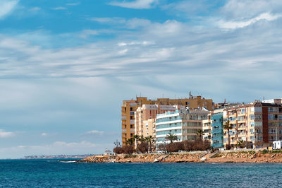 Buildings by sea against cloudy sky