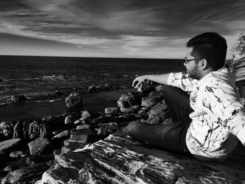 Side view of young man sitting on rock at beach against sky