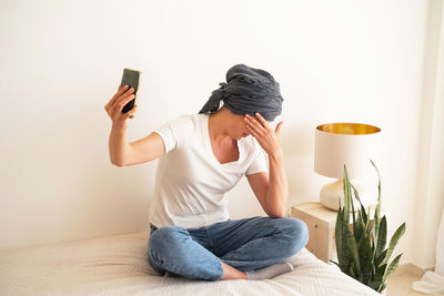 A middle-aged woman sits on a bed looking at a smartphone screen.