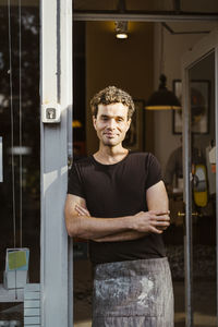 Portrait of smiling male deli owner with arms crossed leaning on doorway