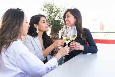 Cheerful adult girlfriends in smart casual clothes sitting at restaurant table and clinking glasses of white wine during meeting on terrace