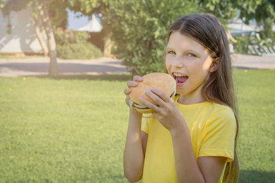 Funny teen girl with a burger in her hands on the background of the park.