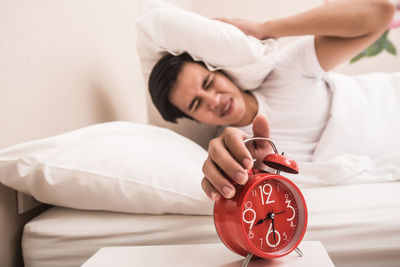 Young man pressing head with pillow while closing alarm clock