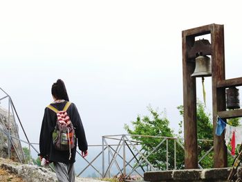 Rear view of woman with backpack walking by bell against clear sky