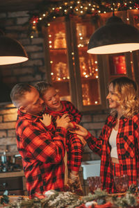 Smile active parents with small son in red checkered sleepwears waiting santa indoor.