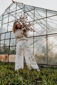 Full length of woman holding plant while standing against greenhouse