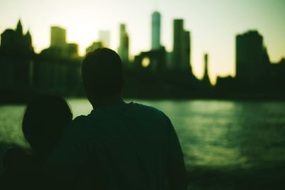 Rear view of man and cityscape against river during sunset