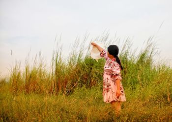 Woman holding hat while standing on field against sky