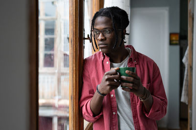 Lonely sad african american man holding mug with tea and looking out window standing indoors