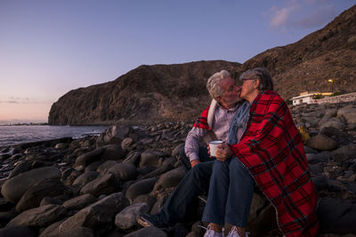 Couple kissing while sitting on rocks against sky at beach