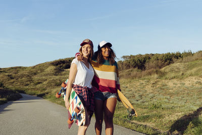Two beautiful multiethnic cheerful women skaters holding skate boards while walking in the middle of a road on a sunny day
