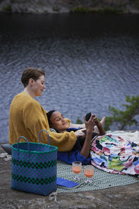 Female couple having picnic by river and using phone