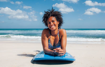 Portrait of smiling young woman lying on surfboard at beach during sunny day