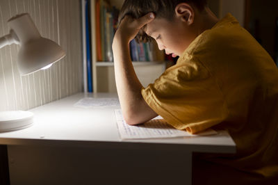 Student with head in hand reading book on table at home