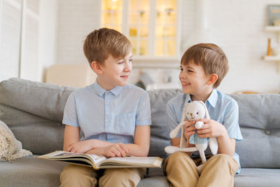 An older brother is reading book to his younger brother sitting on couch.