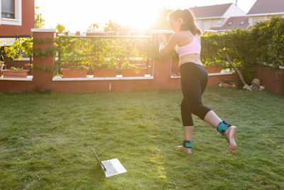 Side view of fit female athlete in sportswear using laptop and doing exercise while following online tutorial during training in backyard at sunset