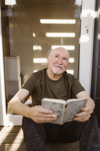 Smiling senior man day dreaming while sitting with book at home