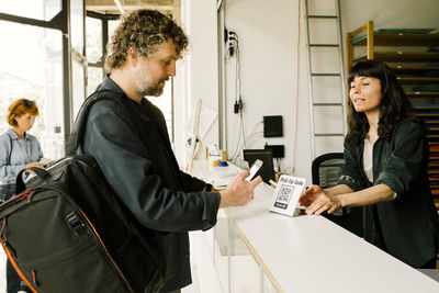 Female technician assisting male customer doing mobile payment at checkout counter in repair shop