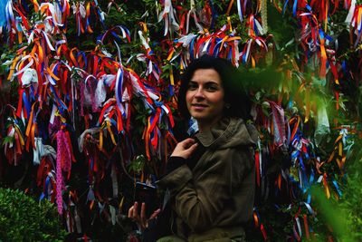 Portrait of smiling young woman standing against colorful ribbons on tree