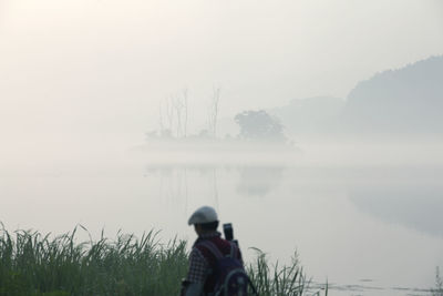 Rear view of man standing at lakeshore during foggy weather