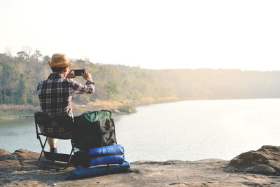 Rear view of man photographing while sitting on chair against river