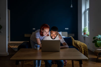 Happy young married multiracial couple using laptop together at home, planning summer vacation