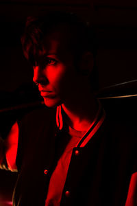 Side view of contemporary female in bomber jacket looking away holding a black baseball bat with red light on background