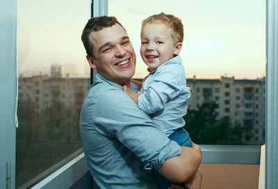 Portrait of happy father carrying son by window
