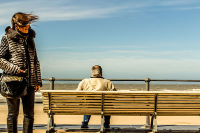 Woman standing by man sitting on bench against sky