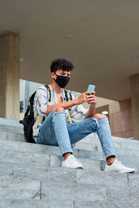 Young afro with mask uses his phone while he is sitting on the stairs