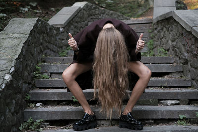 Young woman sitting on steps with upside down head showing thumbs up sign