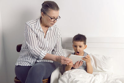 Grandmother in glasses and a white shirt reads a tablet to her grandson lying on the bed in a white 