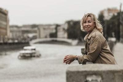 A middle-aged blonde woman with curly hair stands on the river embankment in the city.