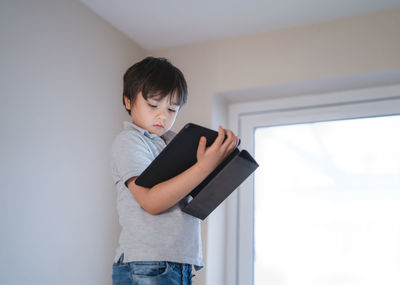 Boy holding mobile phone while standing against wall at home