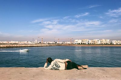 Man sitting on shore by sea against sky in city