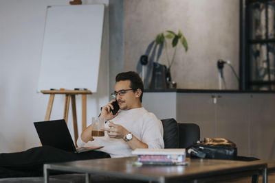 Businessman having coffee while talking on smart phone and working over laptop in office