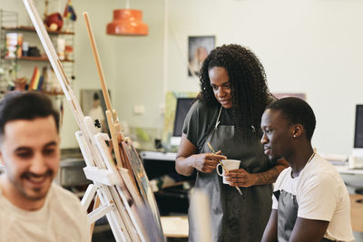 Female tutor looking at painting on easel by male student sitting in art class