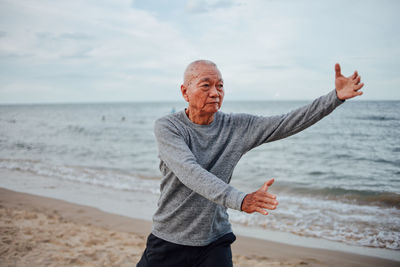 Senior man gesturing while standing on shore at beach against sky
