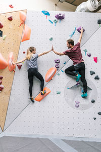 Rear view of male and female friends giving high-five while climbing wall at gym