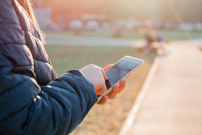 Female hands is holding a smartphone and texting message outdoors in the rays of the evening sun