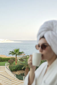 A womaa woman with glasses in a bathrobe and a towel at the hotel drinks coffee from a white cup.