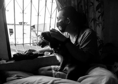 Woman with dog looking through window while sitting on bed at home 