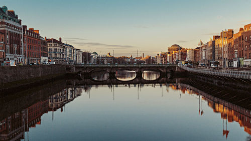 Bridge over river by buildings in city against sky during sunset