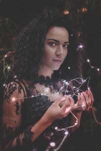 Portrait of young woman holding illuminated string lights by trees at park