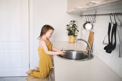 European child of four years old in yellow jumpsuit washes his hands in real bright kitchen