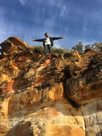 Low angle view of young man with arms outstretched standing on rock against blue sky during sunny day