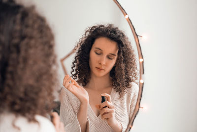 Young woman doing self-care routine at mirror, using aroma oil or serum for skin