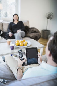 Man holding digital tablet while sitting on sofa with woman in background