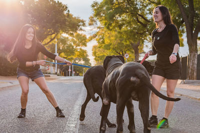 Smiling sisters with dogs standing on road against trees